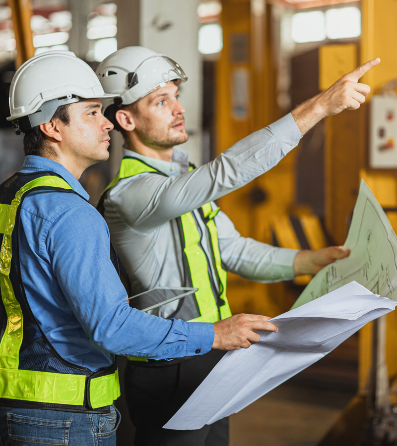 Two male construction workers, including an Electrical Engineering Technician, in safety vests and helmets review blueprints as one points towards something in the distance inside an industrial building or construction site.