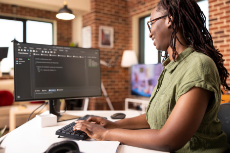An Executive Administrative Assistant with long hair and glasses types on a keyboard at a desk, looking at a computer monitor displaying lines of code in a modern office with exposed brick walls.
