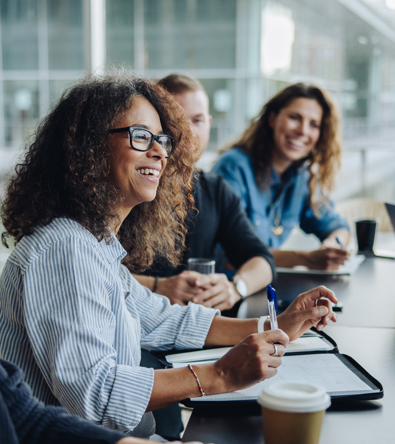 An Executive Administrative Assistant with curly hair and glasses smiles while holding a pen in a meeting with colleagues. They are seated at a table with notebooks, coffee cups, and glass walls in the background.