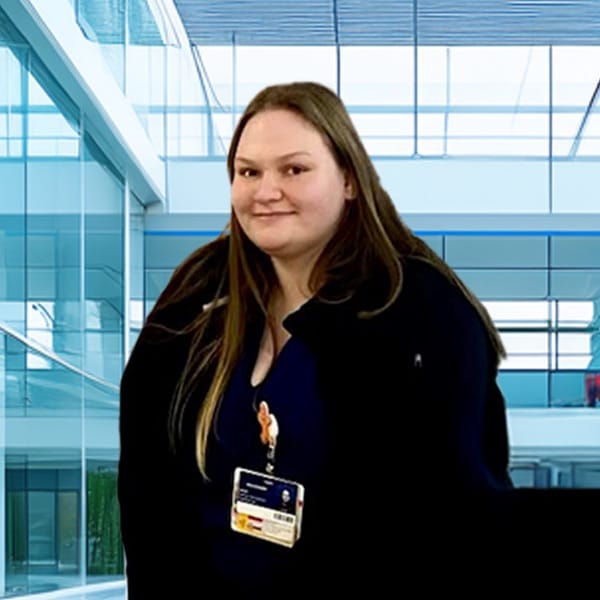 A woman with long brown hair wearing a dark jacket and an ID badge stands indoors in a modern, bright office building with large windows.