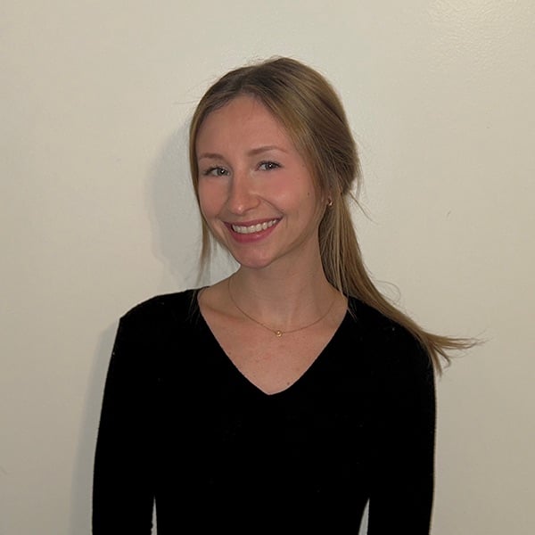 A young woman with light brown hair tied back, wearing a black V-neck top and a delicate necklace, smiles at the camera against a plain light-colored background.