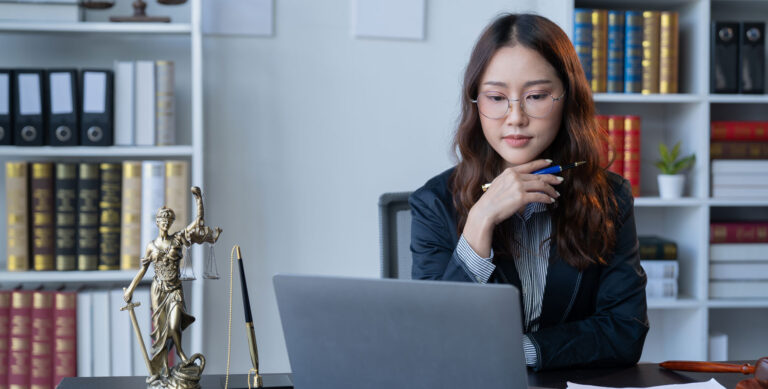 A woman in professional attire sits at a desk with a laptop, holding a pen and looking thoughtful. Law books, a small statue of Lady Justice, and office supplies are visible in the background.
