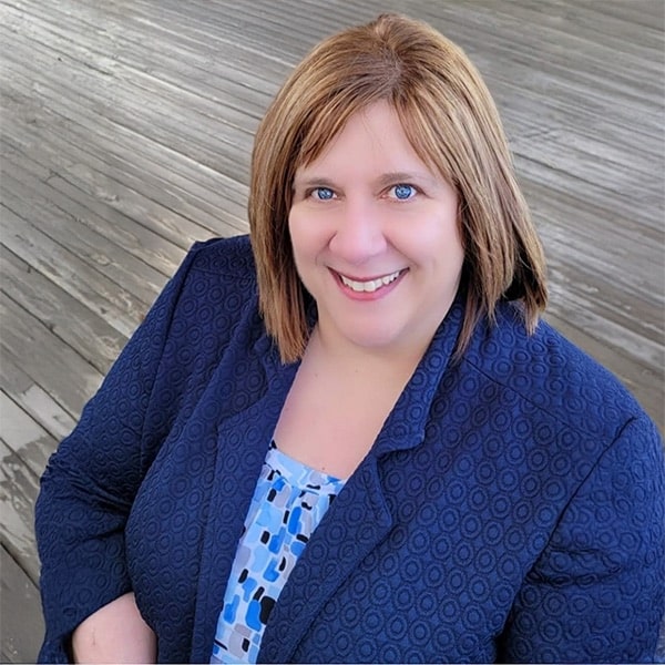 A woman with light brown hair and blue eyes smiles at the camera. She is wearing a dark blue patterned blazer over a blue patterned top, standing in front of a wooden deck background.