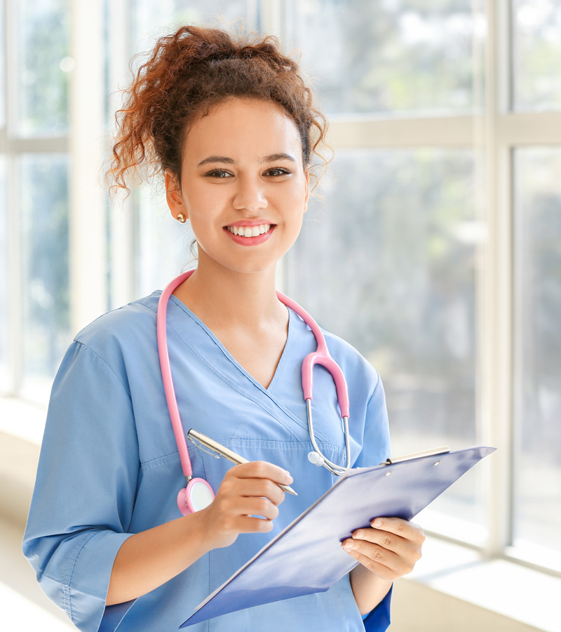 A young Medical Assistant in blue scrubs with a pink stethoscope smiles while writing on a clipboard, standing in a bright, sunlit hallway with large windows behind her.