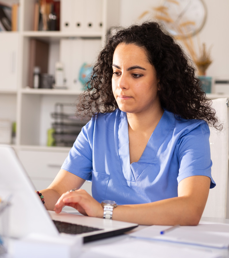 A Medical Assistant in blue scrubs sits at a desk, focused on working on a laptop in a bright office setting with shelves and office supplies in the background.