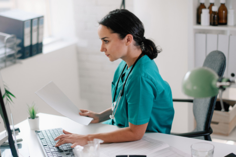 A healthcare worker in teal scrubs sits at a desk, looking at a computer screen while holding paperwork. A stethoscope hangs around her neck as she reviews medical coding documents, with office supplies visible on the desk.