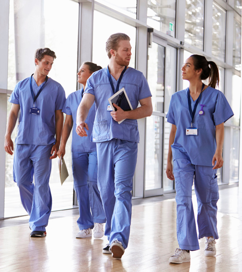 Four healthcare professionals, including a Certified Nursing Assistant in blue scrubs, walk together in a bright hallway, carrying clipboards and tablets while engaged in conversation.