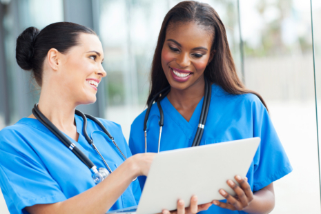 Two female Certified Nursing Assistants in blue scrubs and stethoscopes smile as they look at a tablet together in a bright medical setting.