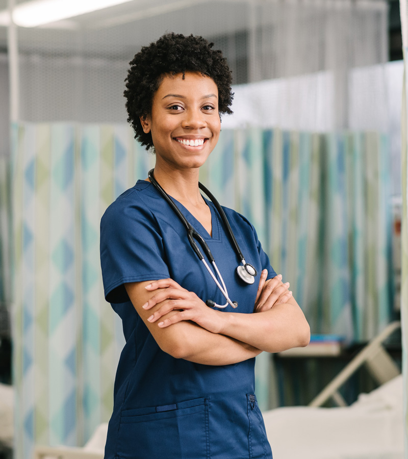 A smiling Certified Nursing Assistant in blue scrubs stands with arms crossed, wearing a stethoscope around her neck, in a medical facility with privacy curtains in the background.