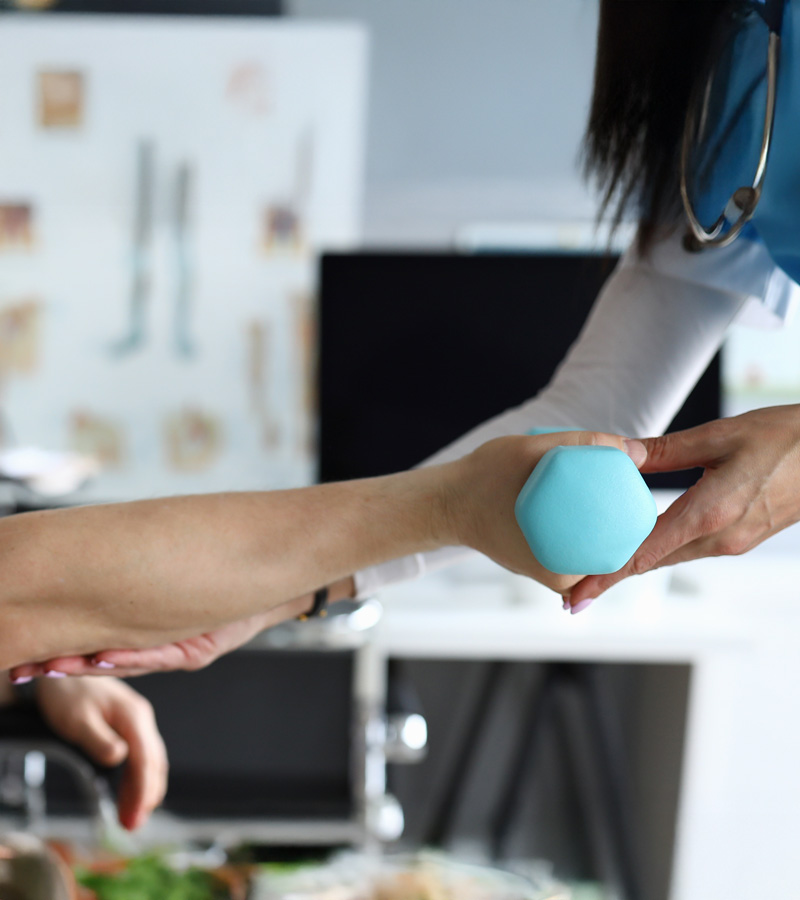 An Occupational Therapy Assistant assists a patient with arm exercises using a light blue dumbbell in a medical office setting.