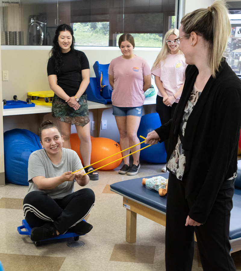 A woman in black holds a yellow exercise band, while another woman, guided by an Occupational Therapy Assistant, sits on a scooter board gripping the band. Three other women stand in the background, watching and smiling in a therapy or classroom setting.