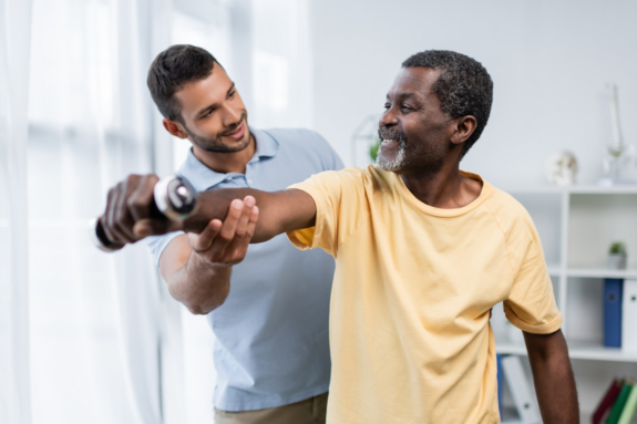 A smiling Occupational Therapy Assistant assists an older man in a yellow shirt with a shoulder exercise, gently supporting his arm in a bright, airy room.