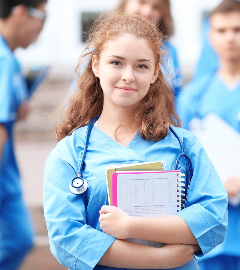 A young woman in blue scrubs, with a stethoscope around her neck, holds notebooks and smiles at the camera—capturing the confidence of a future Patient Care Coordinator. Others in similar attire are blurred in the background, suggesting a medical or nursing school setting.