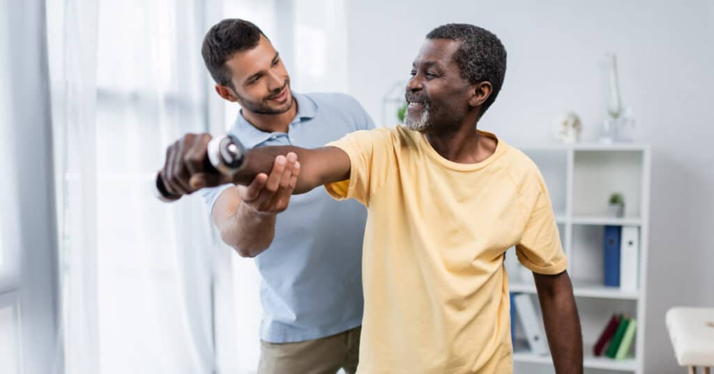 A man in a yellow shirt lifts his arm while a Physical Therapist Assistant encourages and assists him, possibly during a physical therapy session in a bright room.