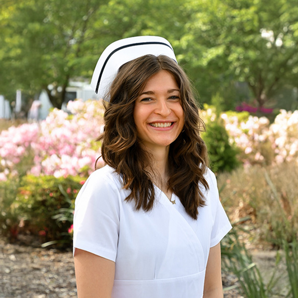 A smiling woman in a white nurse uniform and cap stands outdoors in a garden with blooming flowers and green trees in the background.