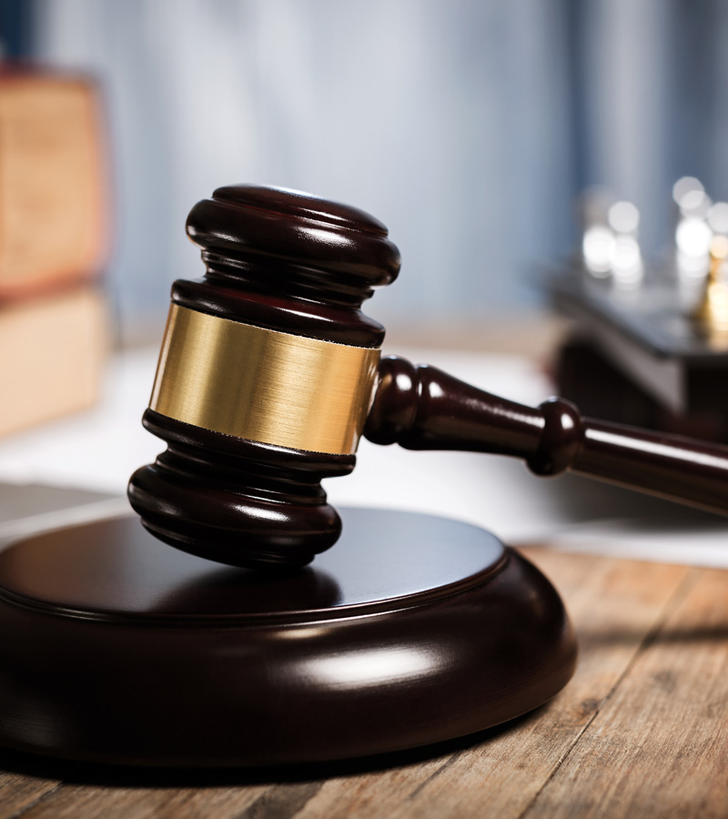 A wooden judge’s gavel with a brass band rests on a sound block on a wooden table, often used by a Sheriff, with blurred legal books and documents in the background.