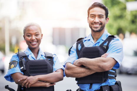 Two Deputy Sheriffs in uniform and black vests stand outdoors with arms crossed, smiling confidently at the camera. The background is slightly blurred, featuring trees and city elements.