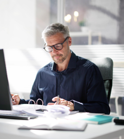 An account clerk with gray hair and glasses sits at a desk in an office, looking down at documents and writing, with a laptop and files in front of him. Sunlight streams through a window in the background as he reviews new account paperwork.