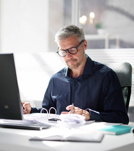 A middle-aged man with gray hair and glasses works at a desk with a laptop, open binder, and papers, concentrating as he writes or reviews documents in a bright office.