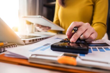 A Budget Analyst in a yellow shirt uses a calculator and holds a tablet, working at a desk with charts, a laptop, documents, and a Budget Template. Sunlight streams in from the left.