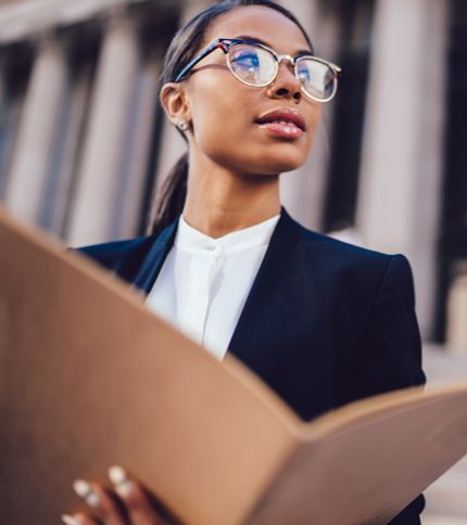 A confident woman wearing glasses and a business suit holds a large folder or document, looking thoughtfully into the distance with a building in the background.