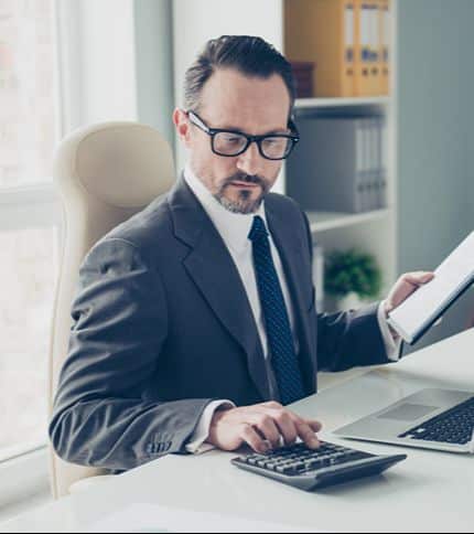 A man in a suit and glasses sits at a desk, using a calculator with one hand and holding papers in the other. A laptop is open in front of him, and shelves with files are visible in the background.