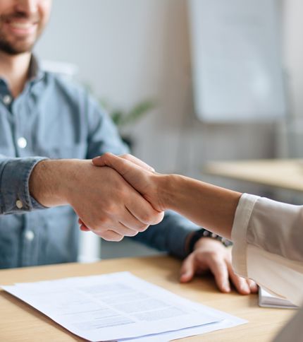 Two people shake hands across a desk with documents on it, suggesting a successful business agreement or interview for a Benefits Coordinator position in an office setting.