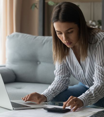 A Billing Clerk sits on a couch, leaning over a table with a laptop and papers, using a calculator and appearing focused on billing cost details.