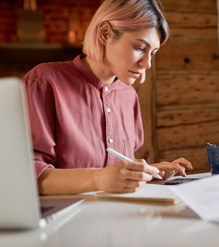A person with short, light pink hair wearing a mauve shirt is sitting at a desk, working on bookkeeping tasks by writing on paper with a pen while looking at a laptop. The background features wooden walls and a soft, warm light.