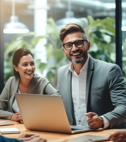 Two professionals, including a Brand Manager, sit at a table with a laptop in an office, smiling and engaged in conversation. The background features green plants and large windows, creating a bright and modern workspace.