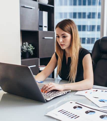 A budget analyst with long hair works at a desk in an office, using a laptop. Papers with charts and graphs are spread out in front of her, alongside shelves stocked with office supplies in the background.