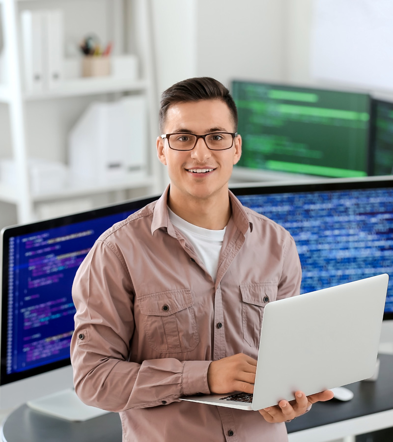 A young man wearing glasses and a light brown shirt is smiling while holding a laptop. He stands in front of two large monitors displaying code, embodying the modern budget analyst at work in a sleek office setting.