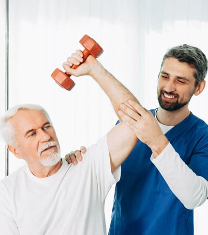 An Occupational Therapy Assistant helps an older man lift a small orange dumbbell during a rehabilitation exercise session. The assistant supports the man's arm and smiles encouragingly.
