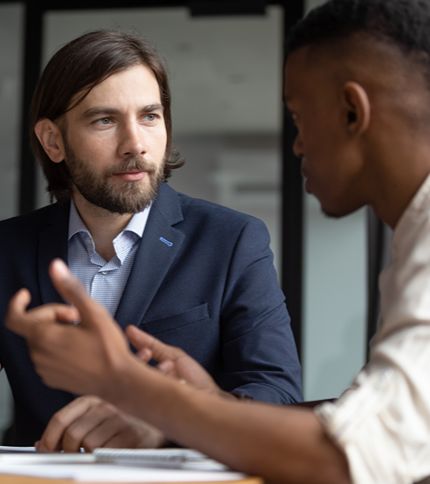 Two men sit at a table having a serious conversation. One, possibly a Case Manager with long hair and a beard, wears a suit jacket, while the other gestures with his hands as he speaks.