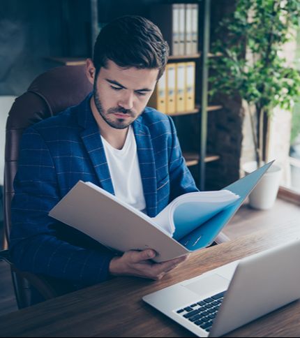 A man in a blue plaid blazer sits at a desk, reviewing documents from a folder. An open laptop—ideal for Case Management Software—is in front of him, with shelves and a tall plant in the background.