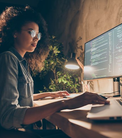 A Coding Support Specialist wearing glasses and a denim jacket sits at a desk, working on code. A lamp, plant, and laptop are nearby, creating a warm, focused atmosphere in the room.
