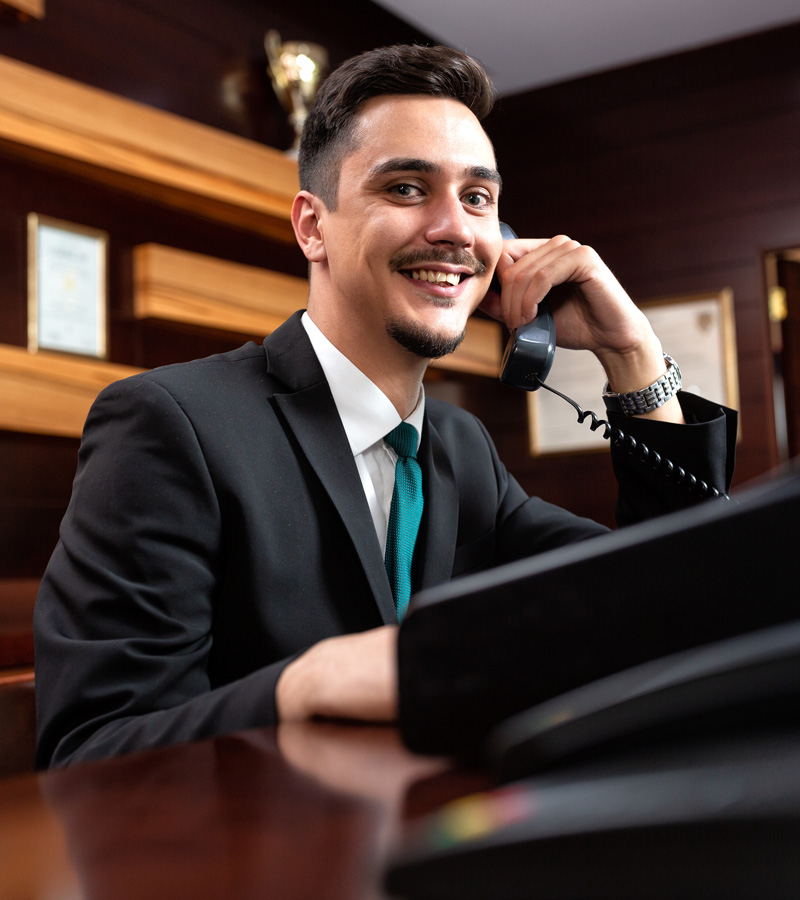 A smiling man in a suit sits at a desk, holding a phone receiver to his ear. He appears to be working at a reception or front desk, embodying the perfect Concierge Template, with certificates and trophies on wooden shelves behind him.