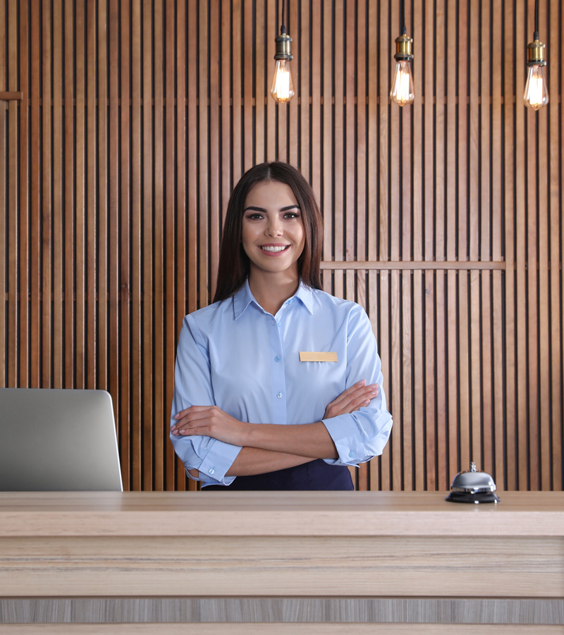 A woman in a light blue shirt stands behind a hotel reception desk, confidently smiling with her arms crossed. A laptop, service bell, and Concierge Template sit on the desk; wooden paneling and hanging lights complete the scene.