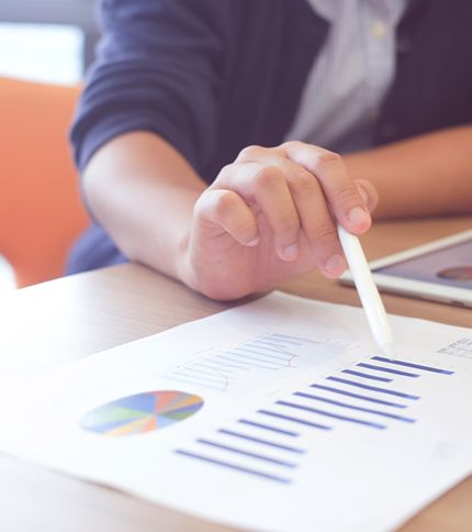 A Data Coordinator points at a printed chart with a pen. The chart features a colorful pie graph and bar graph. The person is sitting at a desk, and a tablet is partially visible next to the paper.