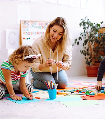 A Daycare Assistant sits on the floor with two young children, helping them draw and play with colored pencils and blocks. Art supplies and colorful papers are scattered around as they enjoy a creative activity together.