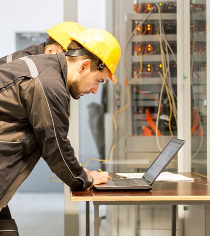 Two Electrical Technicians in yellow hard hats and dark uniforms examine a laptop on a table before a server rack, appearing to check or monitor the system's electrical engineering components.