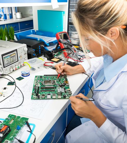 An Electronics Engineering Technician in a lab coat uses a multimeter to test a computer motherboard at a workbench filled with electronic tools, wires, and equipment in a laboratory setting.