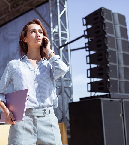 A woman holding a closed laptop and talking on her phone stands outdoors near a stage with large speakers, appearing focused and professional.