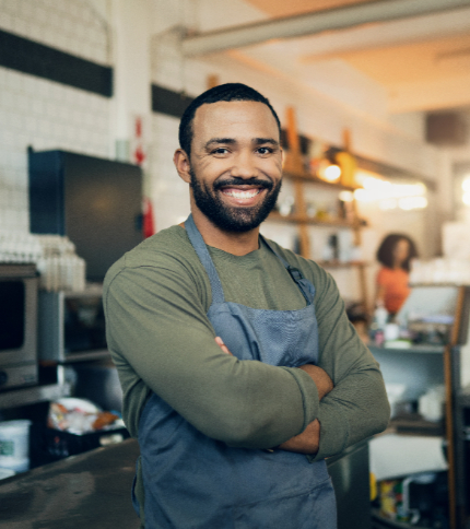 A Food Services Manager wearing a gray apron and green shirt stands with his arms crossed, smiling confidently in a cozy, modern cafe or kitchen. Shelves and various kitchen items are visible in the background.
