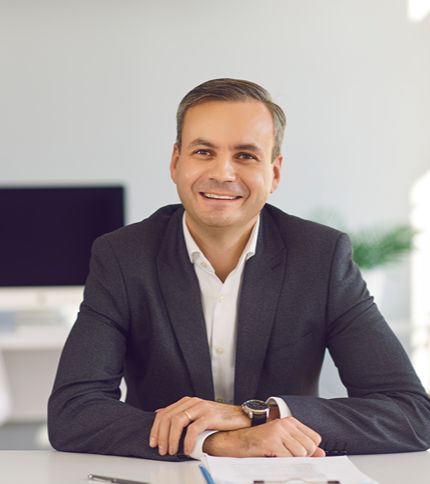 A smiling Operations Manager in a dark suit sits at a desk with documents in front of him. A computer monitor and blurred office decor are visible in the background, creating a modern office atmosphere.
