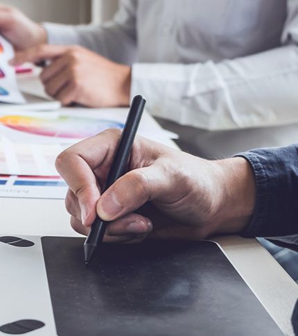 Close-up of a graphic designer’s hand using a stylus on a digital drawing tablet, while another person in the background holds a color chart and works at a desk.