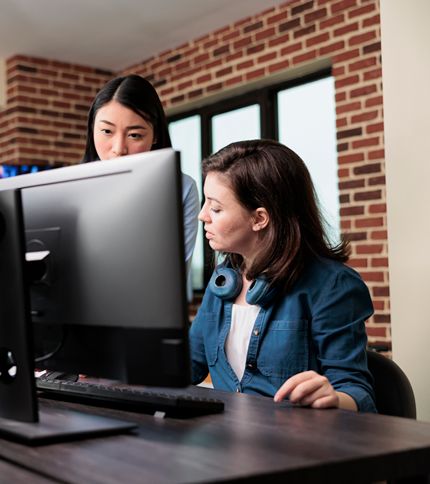 Two women, including a Graphic Production Assistant, work together at a computer in a modern office with brick walls. One is seated at the desk while the other stands beside her, both focused on the monitor.