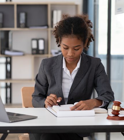 A focused Legal Assistant in a suit sits at a desk, writing in a notebook. A wooden gavel and a laptop are on the desk, and shelves with binders are in the background, highlighting her professional environment.