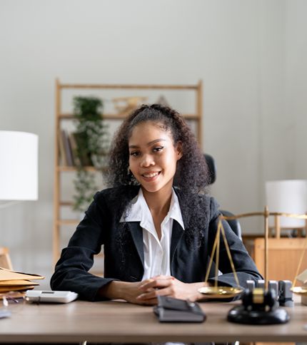 A young legal secretary in a business suit sits at a desk with legal scales, folders, and office supplies, smiling at the camera in a well-lit office with shelves in the background.