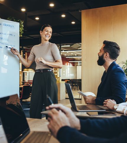 A Marketing Specialist stands at a whiteboard, smiling and presenting to colleagues seated at a conference table with laptops and notebooks in a modern office setting.